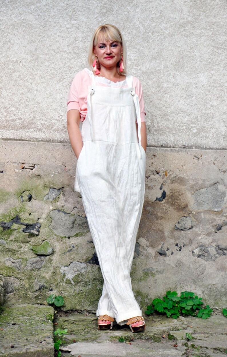 May include: A white linen jumpsuit with adjustable straps and a relaxed fit. The jumpsuit features a bib-style top and wide legs. The model is wearing a pink top underneath and sandals. The background is a textured stone wall.