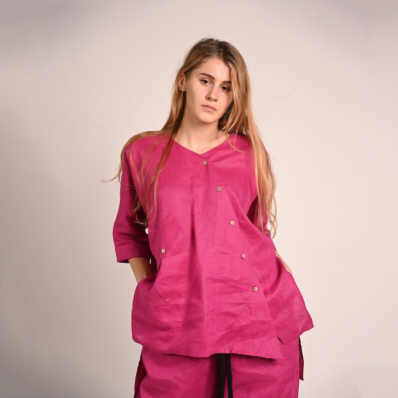 K&ouml;nnte beinhalten: Ein magentafarbenes Leinen-Outfit, bestehend aus einem V-Ausschnitt-Oberteil mit Holzkn&ouml;pfen und Dreiviertel&auml;rmeln, kombiniert mit einer passenden Hose. Das Oberteil hat zwei Vordertaschen. Das Model tr&auml;gt das Outfit.