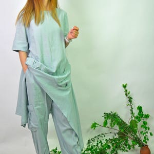 A woman in a light green linen tunic and cargo pants set, with sneakers, stands with one hand near her hip, looking slightly to the left.