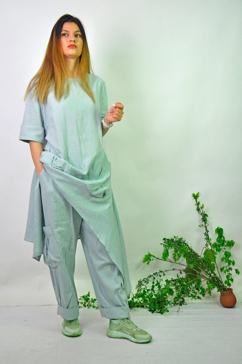 A woman in a light green linen tunic and cargo pants set, with sneakers, stands with one hand near her hip, looking slightly to the left.