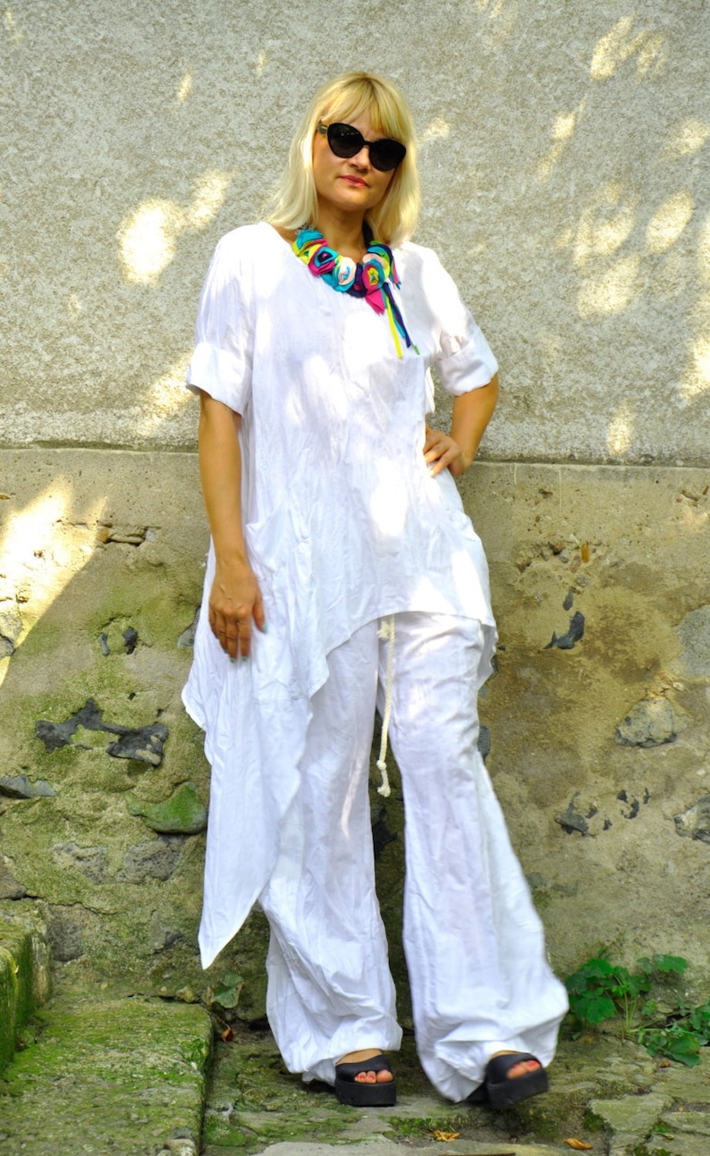 May include: A woman in a white linen outfit, comprising a tunic top and wide-leg trousers. She wears a colourful fabric necklace and black sunglasses. The outfit is completed with black platform sandals.