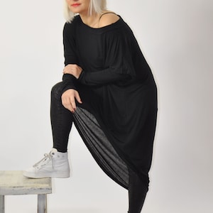 Può includere: Una donna indossa una tunica nera a maniche lunghe con un design drappeggiato e leggings neri. Indossa scarpe da ginnastica bianche e sta in piedi su uno sgabello di legno. Il testo "gabbyga" è in rosa nella parte superiore dell'immagine.