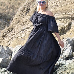 May include: A woman wearing a black off-the-shoulder maxi dress with a tied waist, standing on a rocky cliff. She is wearing blue sunglasses and black platform sandals.