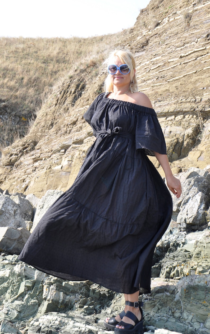 May include: A woman wearing a black off-the-shoulder maxi dress with a tied waist, standing on a rocky cliff. She is wearing blue sunglasses and black platform sandals.