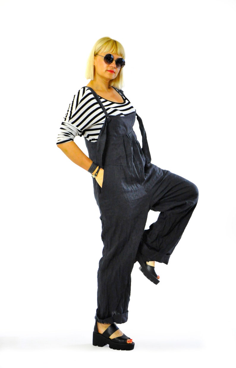 May include: A woman wearing a black jumpsuit with a striped long-sleeve shirt underneath. The jumpsuit has wide legs and suspender straps. She is wearing black platform sandals and sunglasses. The background is white.