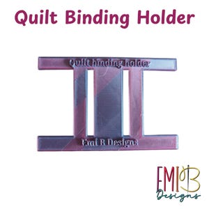 May include: A pink and purple quilt binding holder. The text "Quilt Binding Holder" is at the top and "Emi B Designs" at the bottom. The holder has a rectangular shape with two vertical slots.
