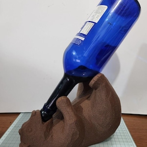 May include: A brown ceramic bear figurine holding a blue glass wine bottle. The bear is lying on its side with its head tilted back, as if it is drinking from the bottle.