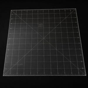 May include: A transparent, square cutting mat with a grid pattern and measurement markings. The mat has diagonal lines and a logo in the center. The edges are marked with centimetre measurements, suitable for crafting and sewing.
