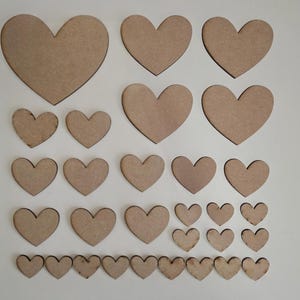 Könnte beinhalten: Eine Sammlung unfertiger Holzherzausschnitte in verschiedenen Größen. Die Herzen sind auf einer weißen Oberfläche angeordnet. Das Holz hat eine natürliche, hellbraune Farbe und die Kanten sind glatt.