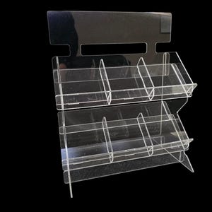 Puede incluir: Expositor de acrílico transparente con dos niveles y múltiples compartimentos. El expositor tiene un cabezal rectangular con un recorte para colgar. Los estantes están inclinados para una óptima visibilidad del producto.