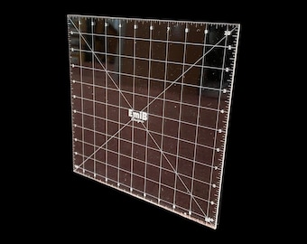 May include: A square, transparent acrylic quilting ruler featuring white grid lines, diagonal markings, and a 10-inch scale along the edges. The center of the tool displays the white text EmiB Designs, providing precise measurements for crafting projects.