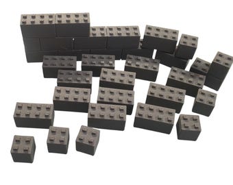 May include: A collection of dark gray interlocking building blocks of various sizes, each featuring a grid of raised circular studs on the top surface. The set includes rectangular and square pieces with a textured finish, arranged in a scattered formation.