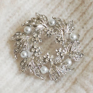 May include: A silver brooch featuring a design of leaves and faux pearls. The brooch is displayed on a white fluffy surface.