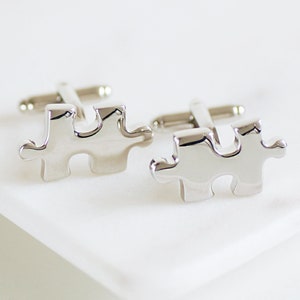 Może przedstawiać: Para srebrnych spinek do mankietów w kształcie puzzli. Spinki do mankietów są wykonane z metalu i mają błyszczące wykończenie.