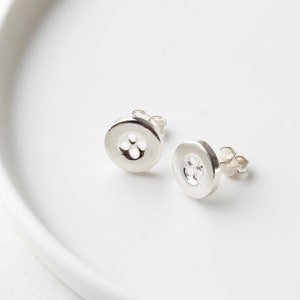 May include: A pair of silver button stud earrings. Each earring has a circular design with three small holes in the center. The earrings are displayed on a white, slightly curved surface.