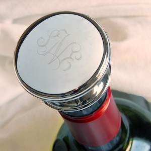 May include: A silver bottle stopper with a monogrammed design. The stopper has a smooth, polished surface and a raised rim.