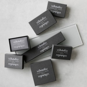 Pode incluir: V&aacute;rias caixas de joias cinza escuro de tamanhos variados, dispostas sobre uma superf&iacute;cie cinza claro. Cada caixa apresenta o texto branco "CARRIAGE TRADE JEWELLERY", com um design floral acima e abaixo do texto. As caixas s&atilde;o para joias.