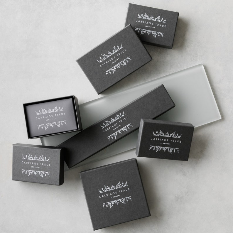 Pode incluir: V&aacute;rias caixas de joias cinza escuro de tamanhos variados, dispostas sobre uma superf&iacute;cie cinza claro. Cada caixa apresenta o texto branco "CARRIAGE TRADE JEWELLERY", com um design floral acima e abaixo do texto. As caixas s&atilde;o para joias.