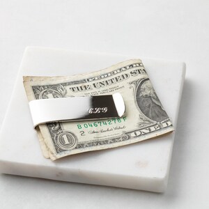 May include: Silver money clip with the initials "CLG" engraved on it. The money clip is holding a US dollar bill.