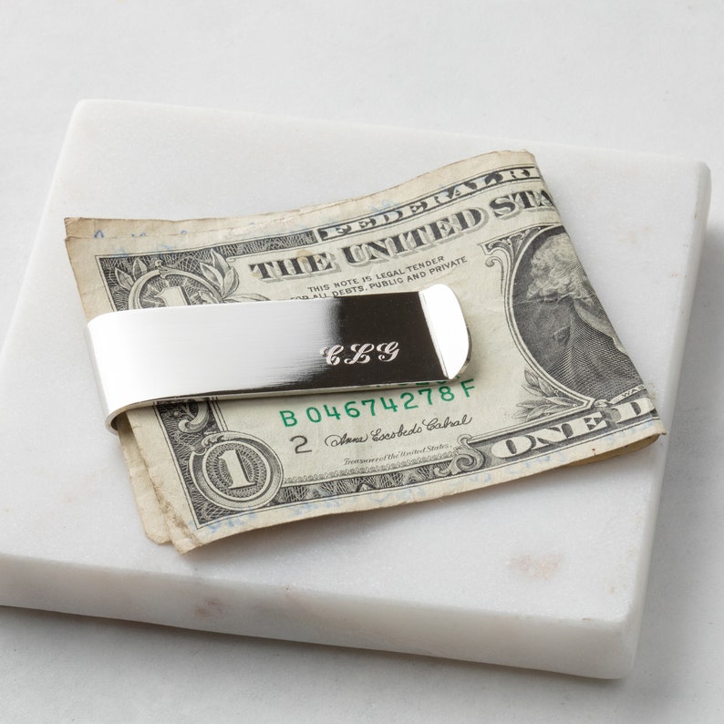 May include: Silver money clip with the initials "CLG" engraved on it. The money clip is holding a US dollar bill with the serial number "B 04674278 F".