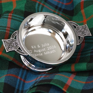 Pode incluir: Uma tigela de prata polida com al&ccedil;as decorativas de n&oacute;s celtas. A tigela tem o texto gravado: "Kit & Julia 22 August 2026 Lainte Mhath". Colocada sobre um tecido xadrez verde, azul e vermelho.