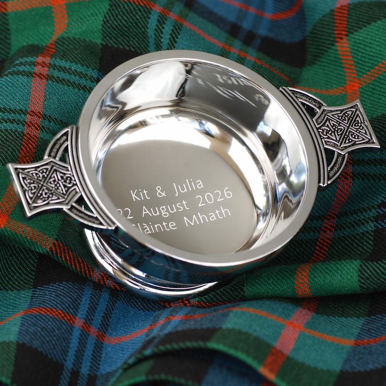 Pode incluir: Uma tigela de prata polida com al&ccedil;as decorativas de n&oacute;s celtas. A tigela tem o texto gravado: "Kit & Julia 22 August 2026 Lainte Mhath". Colocada sobre um tecido xadrez verde, azul e vermelho.