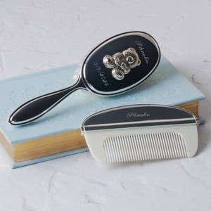 May include: A silver baby brush and comb set. The brush has a black handle and a dark oval head with a teddy bear design. The comb is white with a black top. Both items are engraved with the name "Phoebe".