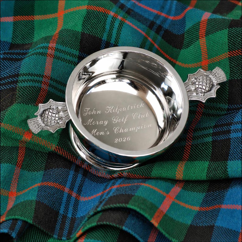 Pode incluir: Uma ta&ccedil;a de trof&eacute;u prateada com a inscri&ccedil;&atilde;o gravada: "John Kilpatrick, Moray Golf Club, Men's Champion, 2026." A ta&ccedil;a tem duas pegas em forma de cardo e assenta num tecido escoc&ecirc;s verde, azul e preto.