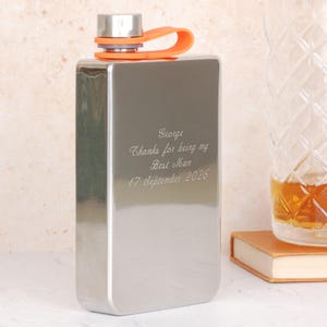Könnte beinhalten: Eine rechteckige Edelstahlflasche mit silbernem Finish, mit einer orangefarbenen Schlaufe und einem silbernen Verschluss. Die Flasche ist mit dem Text "George, Thanks for being my Best Man, 17 September 2026" graviert. Ein dekoratives Glas und ein Buch befinden sich im Hintergrund.
