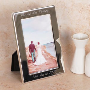 Op de afbeelding: Zilverkleurige fotolijst met de tekst "Mr and Mrs Worthing" en "25th August 2026". De lijst bevat een trouwfoto van een stel. De lijst wordt ondersteund door een zwarte standaard. De achtergrond is neutraal.