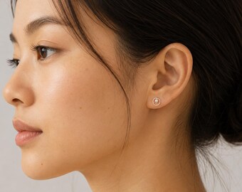 May include: A close-up of an ear with a small stud earring. The earring has a circular design with a small pearl in the centre. The earring is silver in colour and appears to be made of metal.