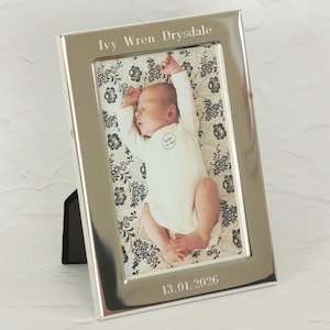 以下が含まれることがあります： 「Ivy Wren Drysdale」の名前と「13.01.2026」の日付が入った銀色のフォトフレーム。 白いロンパースを着た眠っている赤ちゃんが、白黒の花柄の背景に写っている写真が入っています。