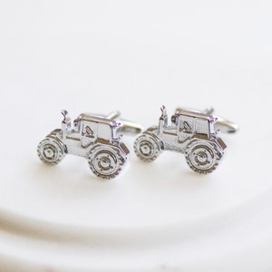 May include: A pair of silver-toned metal cufflinks shaped like tractors. Each cufflink features a detailed side profile of a tractor with a cab, a chimney, and large rear wheels with visible tread patterns, finished with a polished metallic surface.
