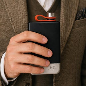 Könnte beinhalten: Eine schwarze Flasche mit silbernem Boden und einem leuchtend orangefarbenen Verschluss. Die Flasche wird von einer Person in der Hand gehalten. Die Initialen "JHK" sind auf dem silbernen Boden eingraviert. Die Flasche ist rechteckig und scheint aus Metall zu sein.