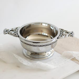 Pode incluir: Uma tigela prateada com al&ccedil;as de n&oacute;s celtas. A tigela tem uma faixa decorativa ao redor do meio e assenta sobre uma base de pedestal. A tigela &eacute; provavelmente feita de metal e &eacute; projetada para servir ou exibir.