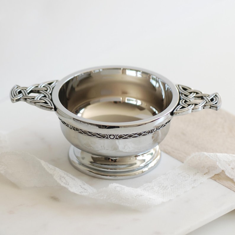 Pode incluir: Uma tigela prateada com al&ccedil;as de n&oacute;s celtas. A tigela tem uma faixa decorativa ao redor do meio e assenta sobre uma base de pedestal. A tigela &eacute; provavelmente feita de metal e &eacute; projetada para servir ou exibir.