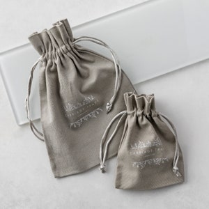 Pode incluir: Duas bolsas cinzentas com cord&atilde;o e cord&otilde;es brancos. As bolsas t&ecirc;m o texto "CARRIAGE TRADE JEWELRY" impresso em branco. As bolsas s&atilde;o feitas de um tecido macio e texturizado e est&atilde;o em tamanhos diferentes.