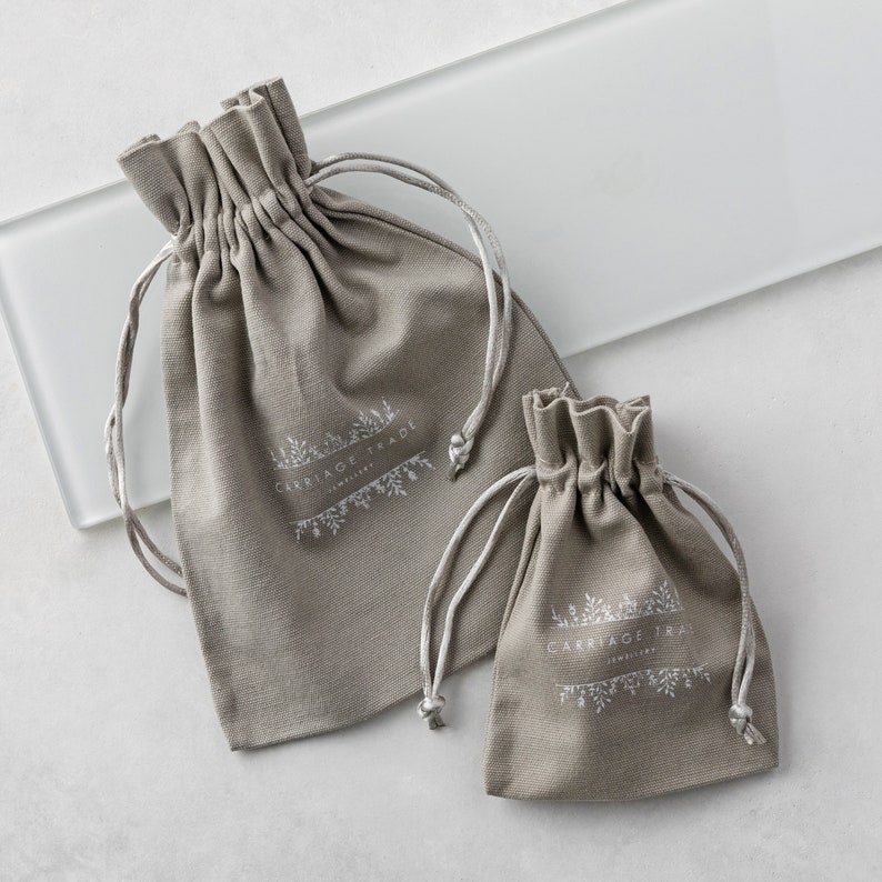 Pode incluir: Duas bolsas cinzentas com cord&atilde;o e cord&otilde;es brancos. As bolsas t&ecirc;m o texto "CARRIAGE TRADE JEWELRY" impresso em branco. As bolsas s&atilde;o feitas de um tecido macio e texturizado e est&atilde;o em tamanhos diferentes.