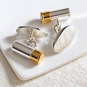 May include: A pair of silver and gold-toned cufflinks shaped like shotgun shell casings. One cufflink features a polished oval face engraved with the cursive initials JHD, while the other displays the cylindrical body and gold-toned base of a shell.