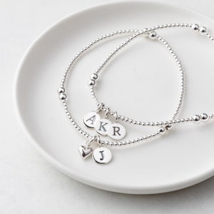 May include: Three silver beaded bracelets with personalised round charms. The charms display the letters A, K, R, and J, along with a heart charm. The bracelets are displayed on a white plate.