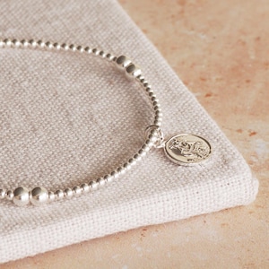 Könnte beinhalten: Ein silbernes Armband mit Perlen und einem silbernen Anhänger mit einer religiösen Figur. Das Armband liegt auf einer hellbraunen Stoffoberfläche.