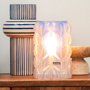 May include: A white, textured glass lamp with a blue iridescent finish. The lamp is lit and casts a warm glow. The lamp is labeled "OHHIO. 3D PRINTED IN LONDON."