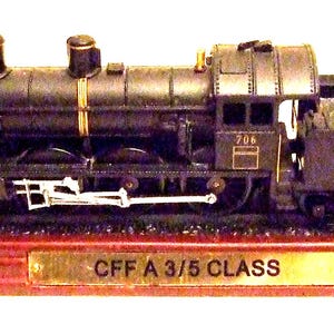 May include: A detailed model train, a CFF A 3/5 Class locomotive, is displayed on a wooden base with miniature tracks. The train is primarily black with gold accents and features intricate detailing. The base has a gold plaque with the text "CFF A 3/5 CLASS".