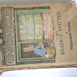May include: Vintage book cover for "Ginger & Pickles" by Beatrix Potter. The cover is tan with the title in a decorative frame. The illustration shows a rabbit in a blue coat looking out a window.