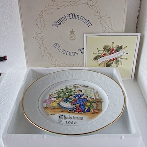 May include: A white porcelain plate with a gold rim featuring a Christmas scene with children around a decorated Christmas tree. The plate is in a white box with a Royal Worcester Christmas Plate 1980 label.