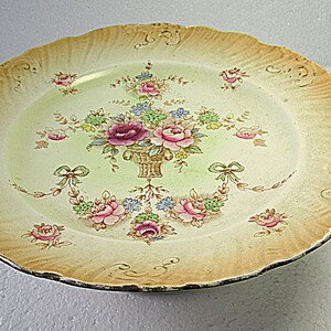 May include: A round ceramic plate with a scalloped edge, featuring a pale green center decorated with a floral basket motif in pink, blue, and green. The wide, textured rim is shaded in a gradient of light orange and tan with embossed details.
