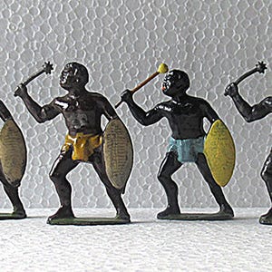 May include: Four black warrior figurines, each with a weapon and shield. The warriors wear loincloths in blue, yellow, and red. The figurines stand on a green base. The background is textured.