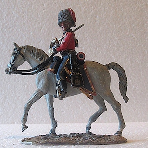 Może przedstawiać: Miniaturowa figurka żołnierza na koniu. Żołnierz ma na sobie czerwoną kurtkę, ciemnoniebieskie spodnie i wysoki czarny kapelusz. Koń jest szary z czarną uzdą i siodłem. Figurka stoi na małej, brązowej podstawie.