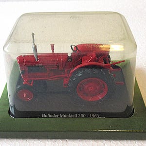 May include: A red Bolinder Munktell 350-1963 model tractor displayed in a clear plastic case. The miniature tractor has black tires and is set on a green base. The text "Bolinder Munktell 350 - 1963" is printed on the base.