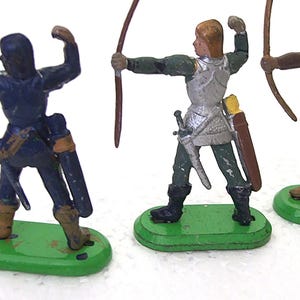 Op de afbeelding: Drie speelgoedboogschutters in verschillende kleuren, elk met een getrokken boog. De figuren staan op groene bases. Eén is blauw, één is zilver en groen, en de andere is groen en zwart. Elk heeft een koker en een zwaard.